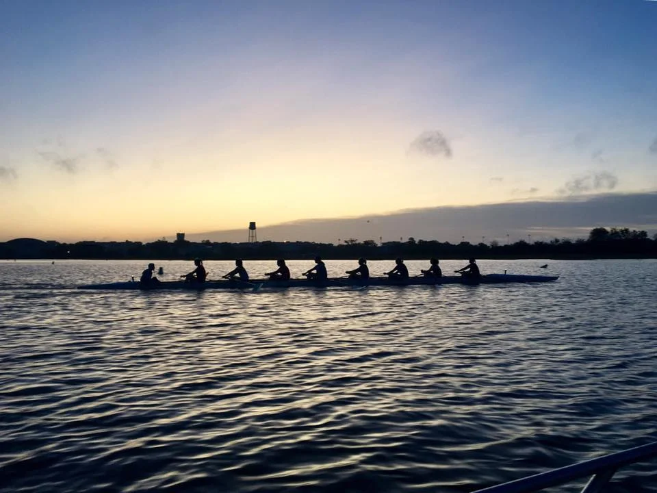 Silhouette of a long rowing crew boat with multiple rowers gliding across calm water at dusk, with a water tower and low buildings on the horizon in muted blue and gray tones