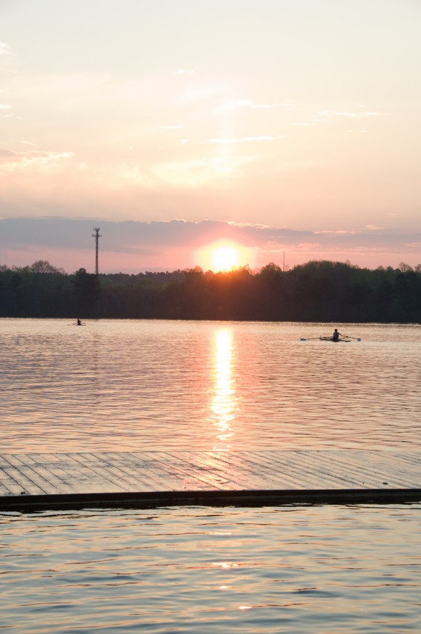 Sunset over a calm lake with two small rowing boats visible in silhouette, warm orange and pink sky tones, tranquil athletic mood.